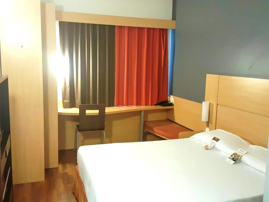 Quarto pequeno com cama de casal com lençol branco, cabeceira de madeira, mesa e cadeira ao lado da cama e cortina marrom e vermelha. Imagem para ilustrar o post hotéis e pousadas em Itu.