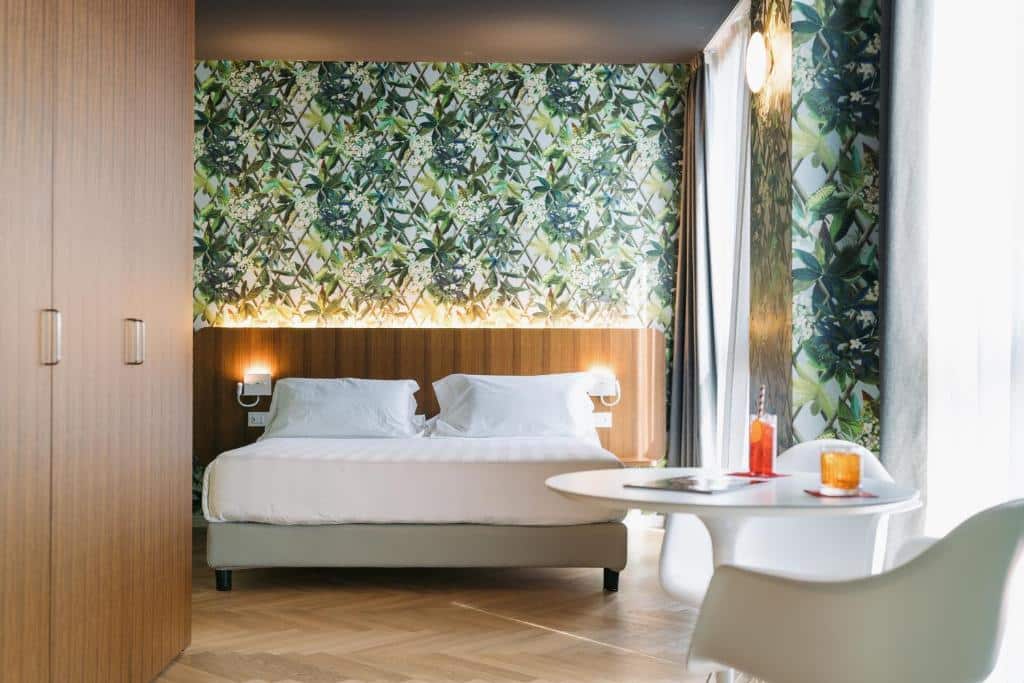 Quarto do Collini Rooms com uma cama de casal, um armário amplo, uma cabeceira com luminárias presas, duas janelas com cortinas e uma mesinha redonda com suas cadeiras, para representar os hotéis românticos em Milão