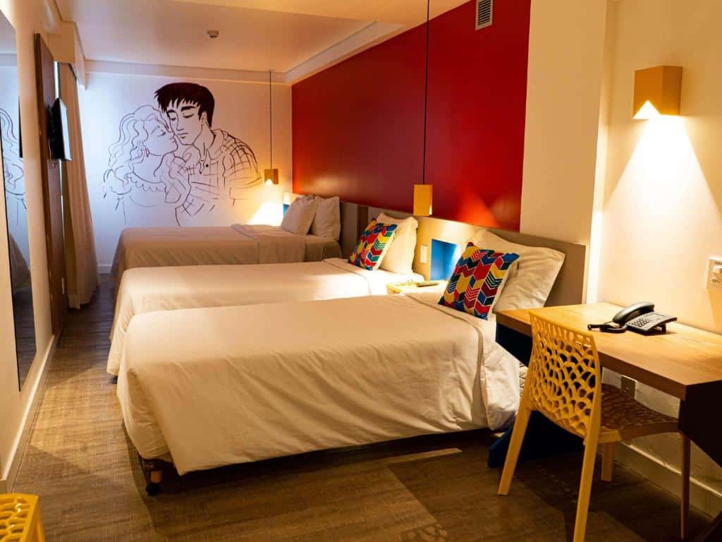 Quarto de hotel com decoração temática junina e nordestina, três camas de solteiro, uma parede vermelha, três luminárias, mesa de madeira e cadeira amarela. Imagem para ilustrar o post hotéis em Campina Grande.
