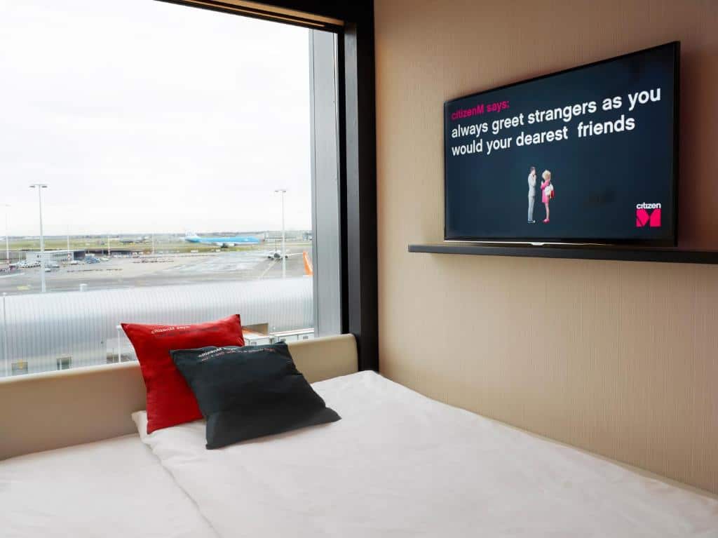 Parte da cama de um dos quartos do citizenM Schiphol Airport, com dois travesseiros (um vermelhor e outro preto) em cima e uma TV ligada ao lado suspensa na parede. Ao lado esquerdo, há uma janela de vidro com vista do aeroporto. Imagem para ilustrar o post de hotéis em Amsterdam perto do aeroporto