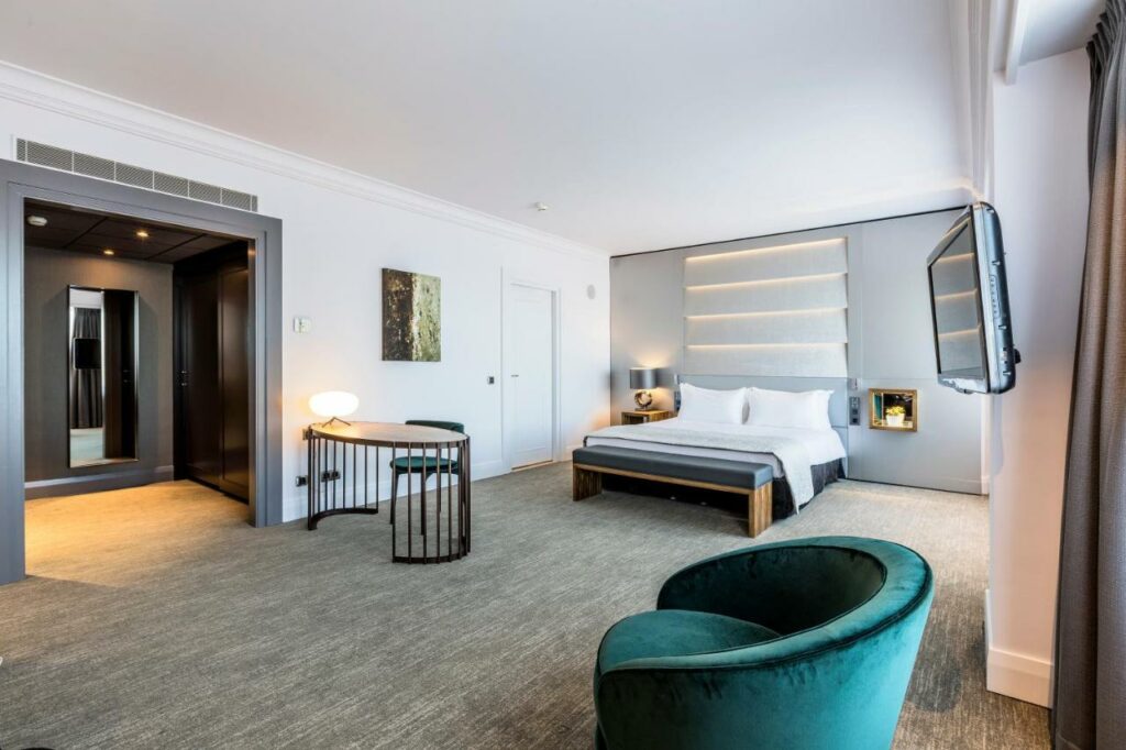 Quarto do Hotel Okura Amsterdam, com uma cama de casal, uma poltrona estofada verde e uma mesa com madeira. O ambiente é espaçoso e tem bastante espaço entre um móvel e outro
