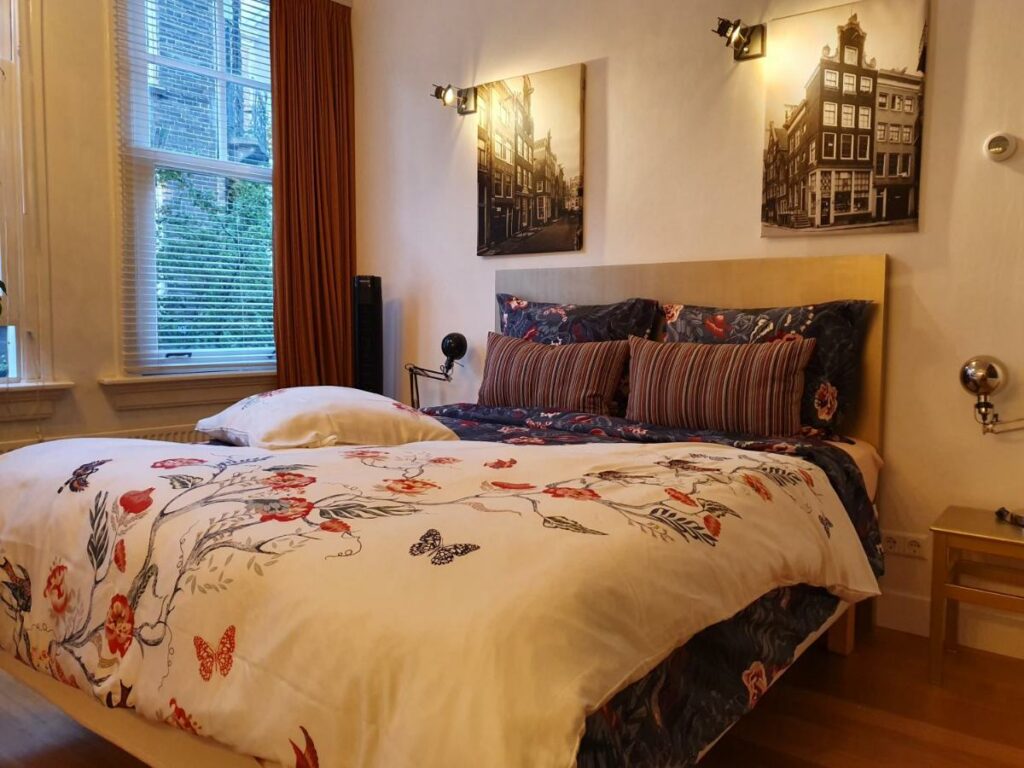 Quarto do apartamento Huis Roomolen, um dos aluguel de temporada em Amsterdam, com uma cama de casal macia com roupa de cama com desenhos de borboletas e flores, além de travesseiros em cima da cama. Há dois quadros com uma luz ao lado de cada iluminando desenhos preto e branco de prédios históricos, além de uma janela no lado esquerdo com uma cortina vermelha recolhida ao lado