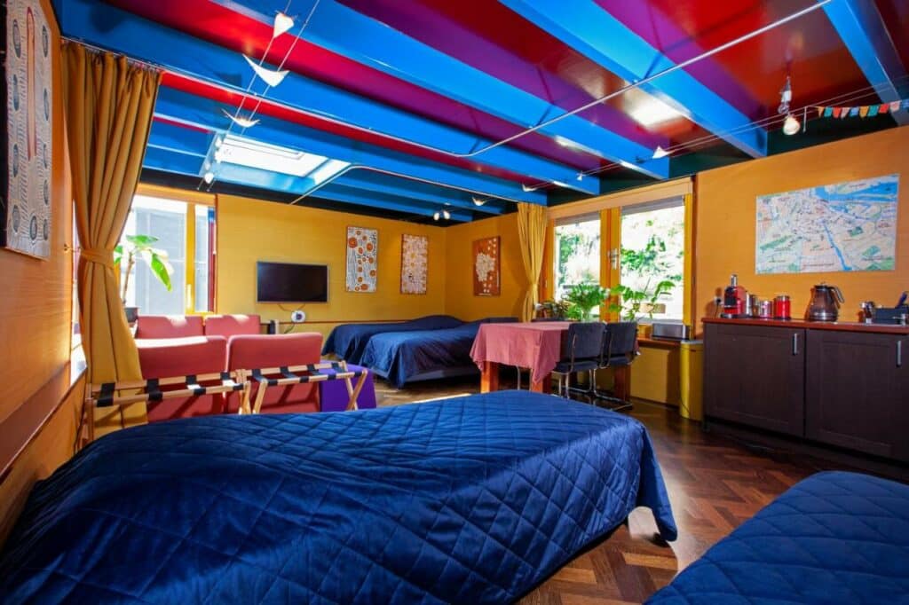 Interior do Dreamtime Houseboat, com 4 camas de solteiro com colchas azuis escuro, duas mesas com 4 cadeiras, uma TV suspensa, um balcão de madeira com amenidades de cozinha e duas janelas com vista da natureza. As paredes do local são amarelas e o teto vermelho e azul