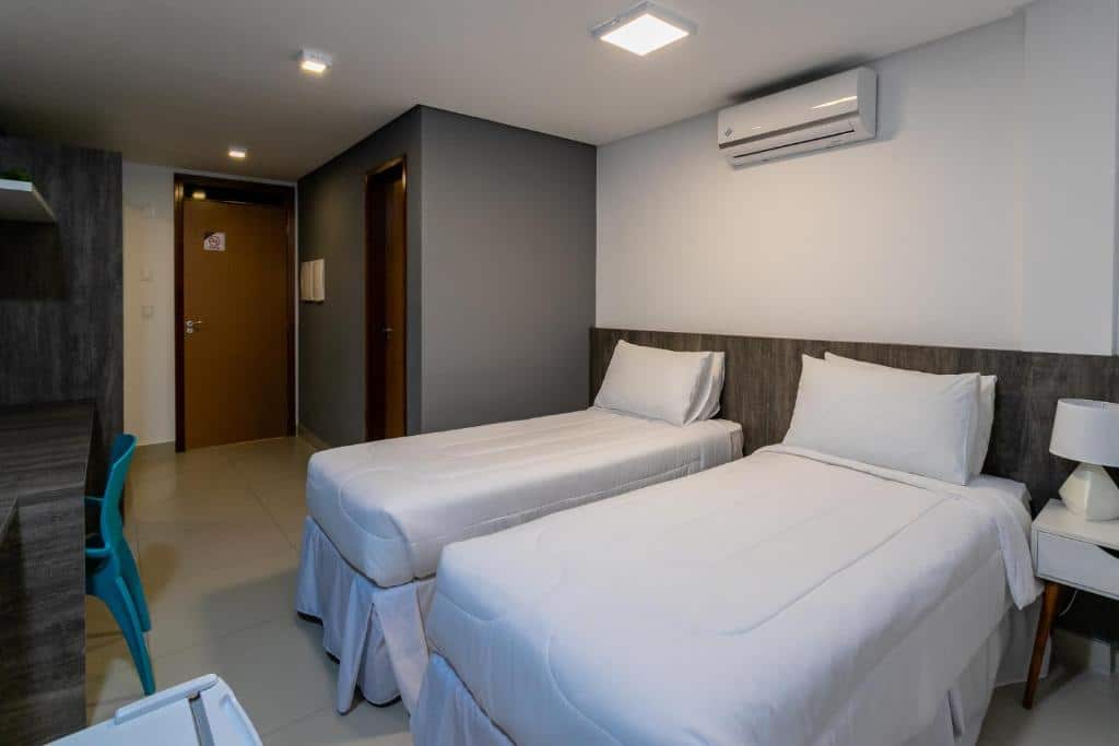 Quarto de hotel simples com duas camas de solteiro, ar-condicionado em cima da cama, cabeceira de madeira, uma parede cinza e móveis de madeira. Imagem para ilustrar o post hotéis em Campina Grande.
