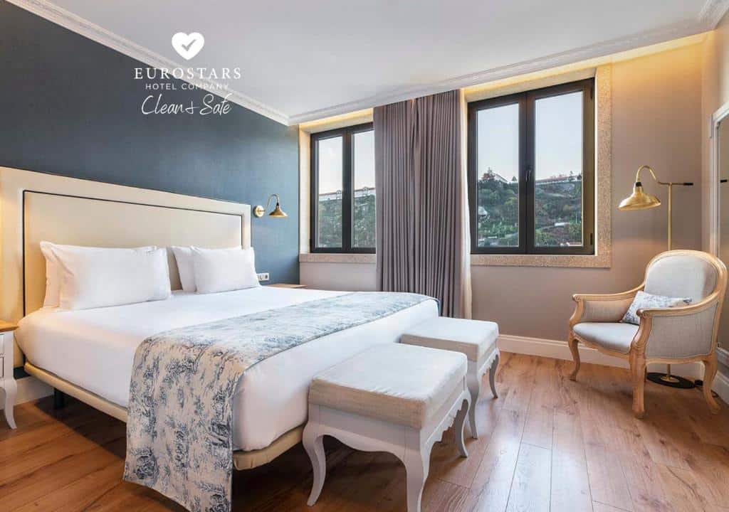 Quarto do Eurostars Porto Douro com cama de casal do lado esquerdo da imagem, no pé da cama dois bancos estofados e em frente a cama uma poltrona. Representa hotéis bem localizados no Porto.