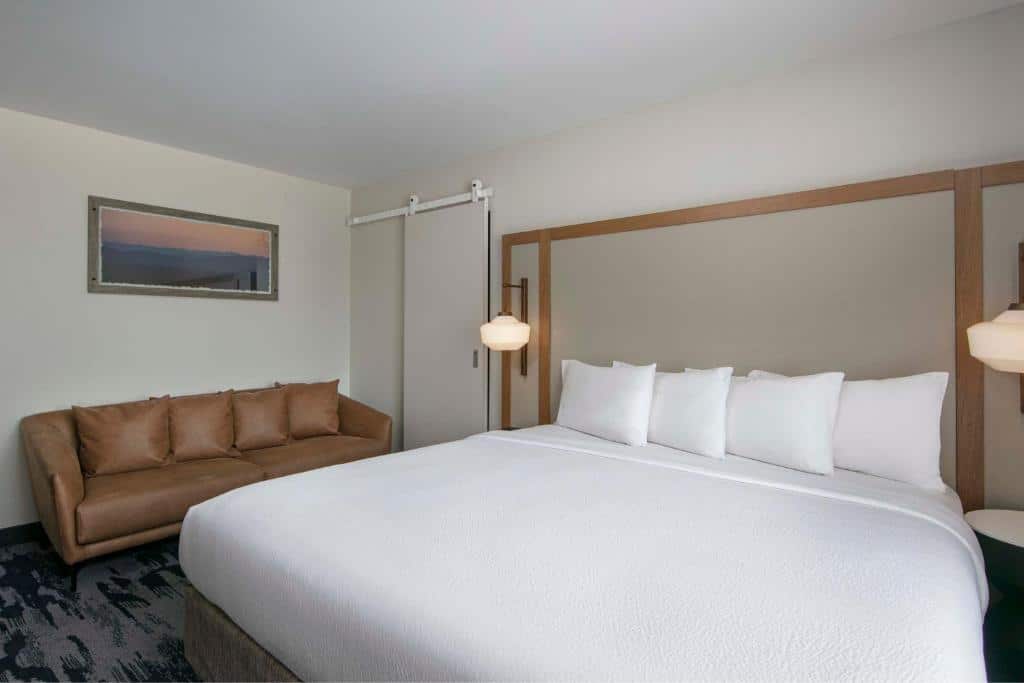suíte do Fairfield Inn by Marriott com uma cama de casal com quatro travesseiro brancos e um sofá de quatro lugares em tom bege ao lado da cama.