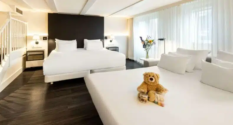 Quarto família do NH Collection Amsterdam Barbizon Palace, de 30 m², com duas camas de casal, sendo uma delas com um ursinho em cima e alguns mimos para crianças, e entre as camas tem uma mesa redonda com um vaso de flores em cima. Representa hotéis para família em Amsterdam