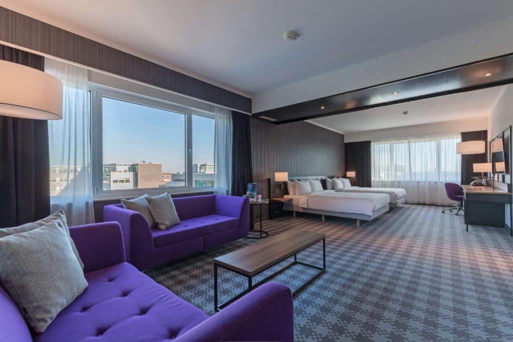 Quarto família do Radisson Blu Hotel Amsterdam Airport, de 49 m², com área de estar com dois sofás roxos, uma mesa ao centro e, no lado esquedo, há duas camas de casal e uma mesa de trabalho a frente. Há janelas com vista da cidade