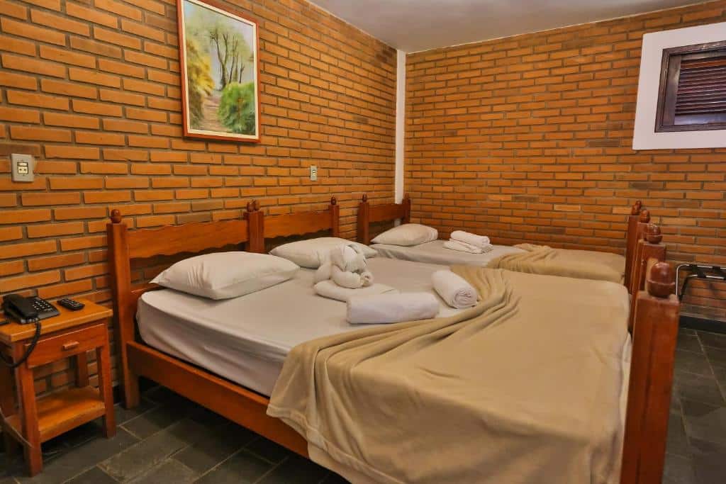 Imagem do quarto do Hotel Fazenda Campo dos Sonhos, em Socorro, para ilustrar o post sobre pousadas perto de Ribeirão Preto. As paredes são de tijolos, e as camas de madeira. Na suíte, há uma cama de casal e outra de solteiro. Toalhas de banho estão dispostas em cima delas.