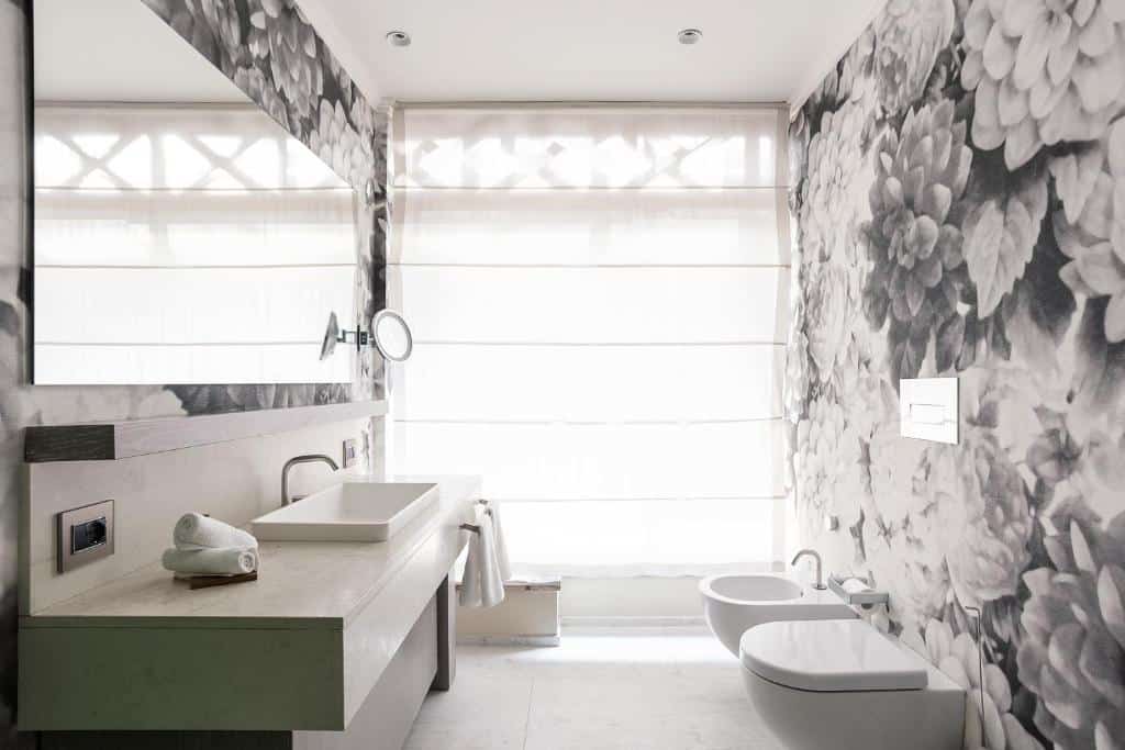 Banheiro espaçoso do Galleria Vik Milano com uma pia ampla com toalhas brancas, um grande espelho, um vaso e um bidê, o local tem papéis de parede com rosas pretas e brancas desenhadas