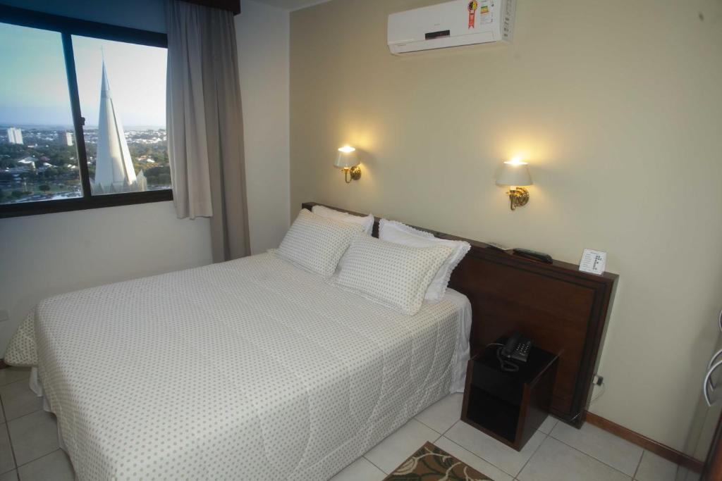 Quarto do hotel com uma cama de casal branca, móveis de madeira, duas lâmpadas na parede e uma janela de vidro com cortina com vista para a paisagem da cidade.