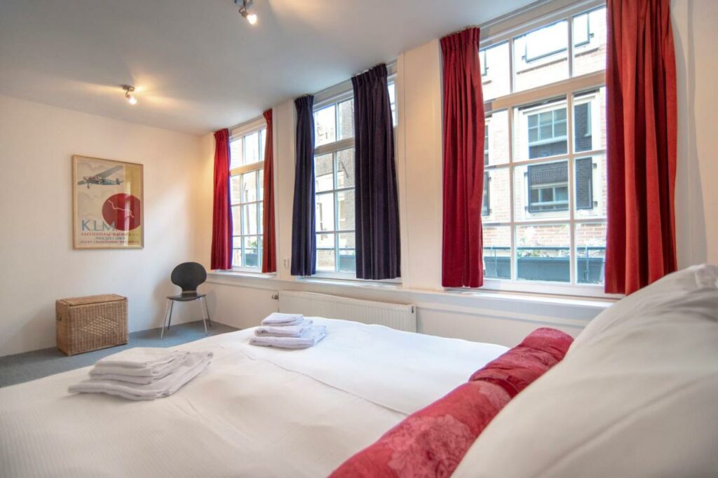 Cama de casal com toalha dobrada em cima, uma cadeira e um baú na frente e no lado direito tem janelas com cortinas pretas e vermelhas. Representa airbnb em Amsterdam