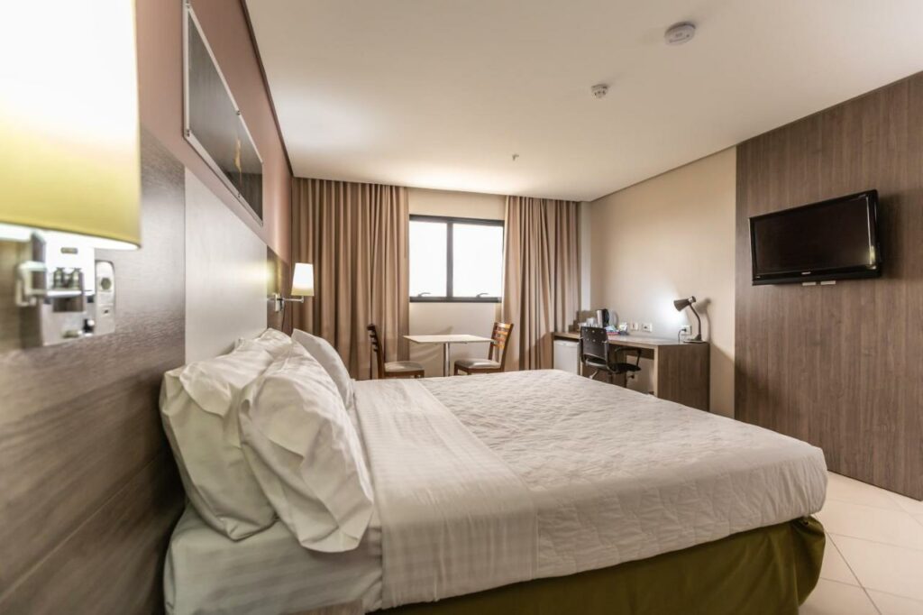 Quarto do hotel Holiday Inn Cuiabá com uma cama de casal, janela com cortinas, mesa de trabalho com cadeira e luminária, mesa para duas pessoas e uma televisão. Foto para ilustrar post sobre hotéis em Cuiabá.