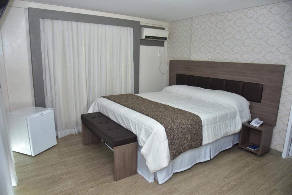 Quarto de hotel espaçoso com cama de casal com colcha branca e detalhe marrom, cabeceira marrom, cortina grande branca, frigobar e ar-condicionado. Imagem para ilustrar o post hotéis em Campina Grande.