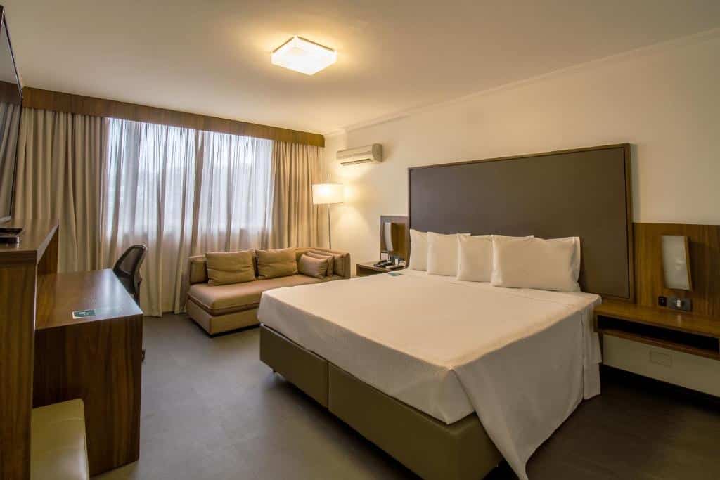 Quarto do hotel com uma cama de casal branca, um sofá pequeno, uma mesa com cadeira e uma janela com cortina.