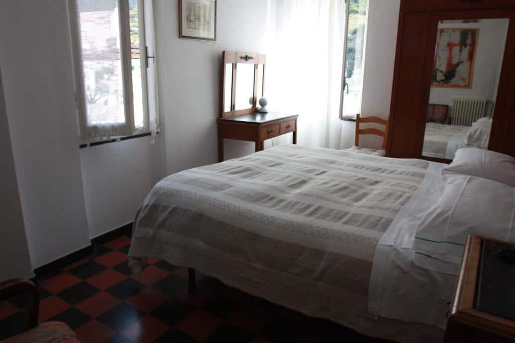 Quarto do hotel com uma cama de casal branca, chão quadriculado, penteadeira, cadeira e guarda-roupa de madeira, paredes brancas e duas janelas de vidro.