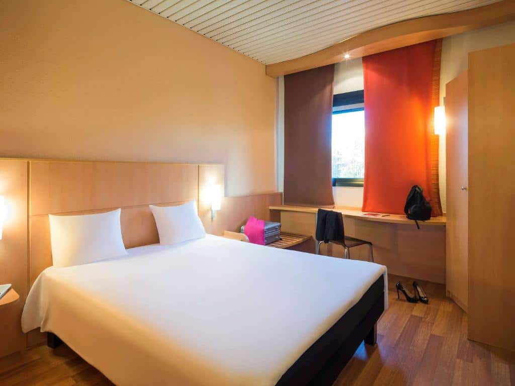 Quarto do Hotel Ibis Milano Ca’ Granda com uma janela com cortinas, uma bancada de escritório com uma cadeira, uma cama de casal e um pequeno armário, para representar hotéis Mercure em Milão