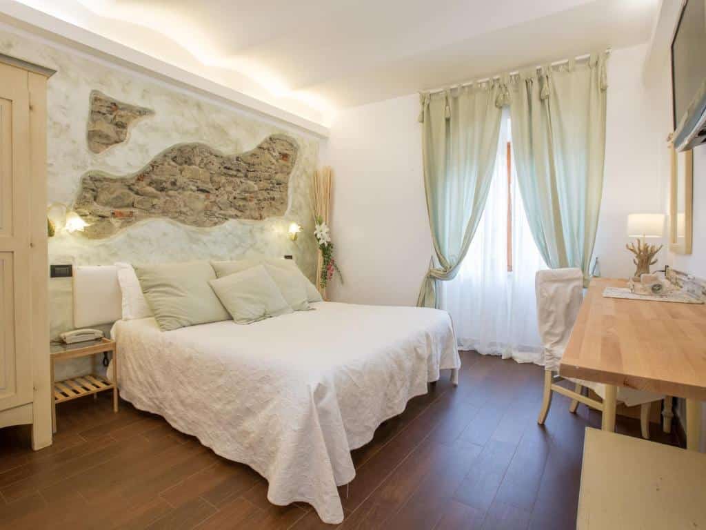 Quarto com uma cama de casal branca e vários travesseiros, chão de madeira, mesa de madeira com uma cadeira, janela com cortina e parede com detalhes de pedras.
