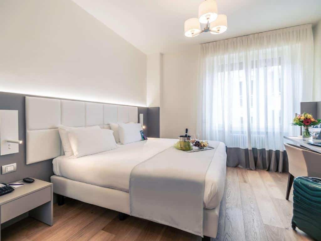 Quarto do Hotel Mercure Milano Solari com uma cama de casal, sob ela há uma bandeja com frutas e um champanhe, há uma janela com cortinas e uma mesa de escritório com uma cadeira, para representar hotéis Mercure em Milão