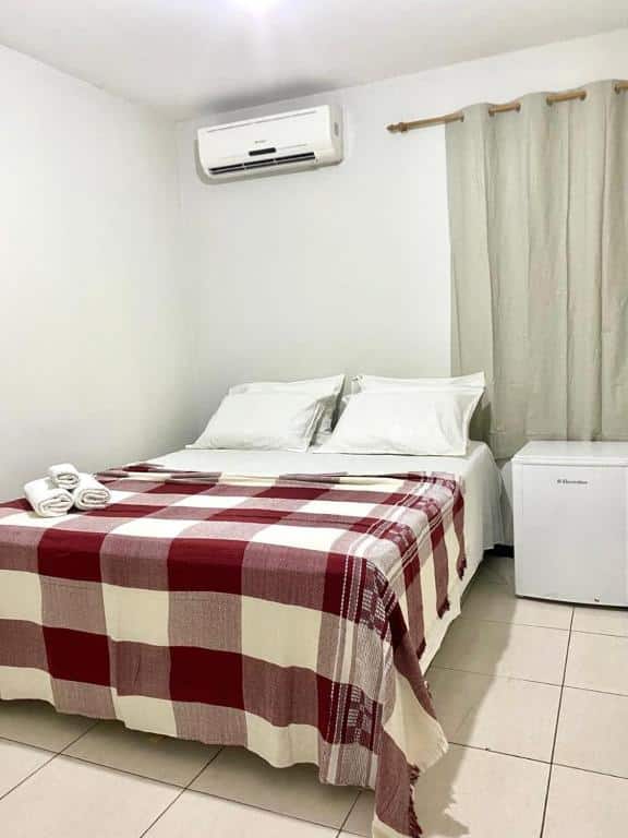Quarto de hotel simples com paredes brancas, cama com colcha branca e xadrez, ar-condicionado, cortina beje e frigobar. Imagem para ilustrar o post hotéis em Campina Grande.