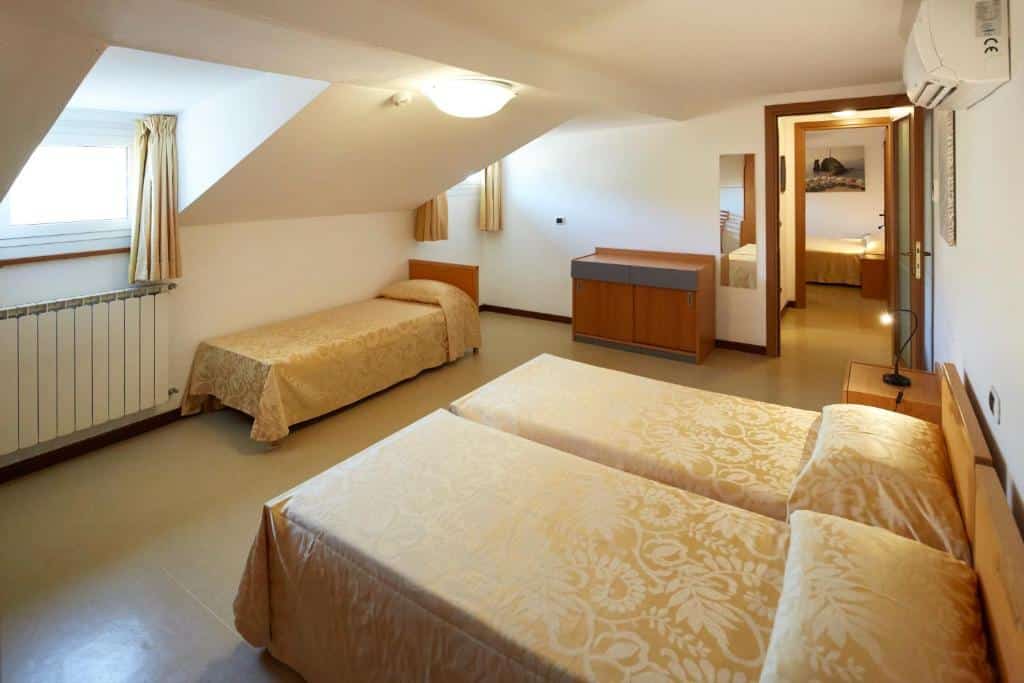 Quarto do hotel com três camas, móveis em madeira, paredes brancas e ao fundo um corredor que dá acesso a mais um quarto.