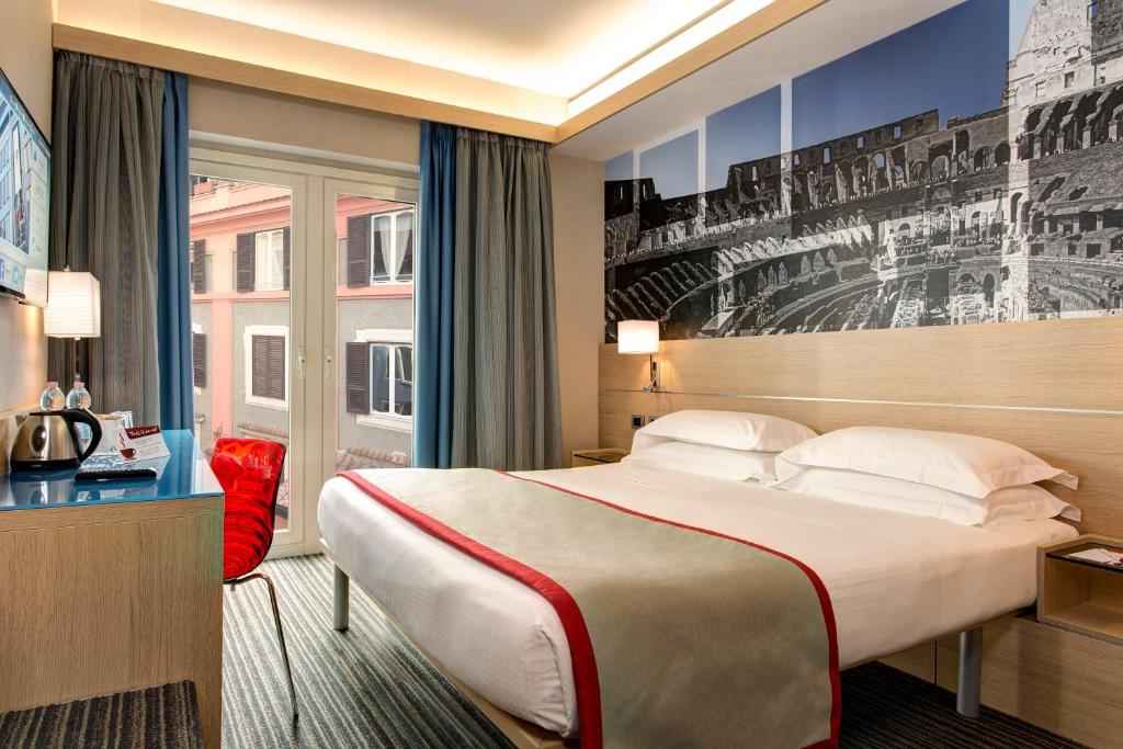quarto do iQ Hotel Roma, um dos hotéis para brasileiros em Roma, com cama de casal, mesinha e luminária em ambos os lados, painel grande com vista do Coliseu acima, mesa com cadeira à frente e janela grande com cortinas, os detalhes são vermelhos