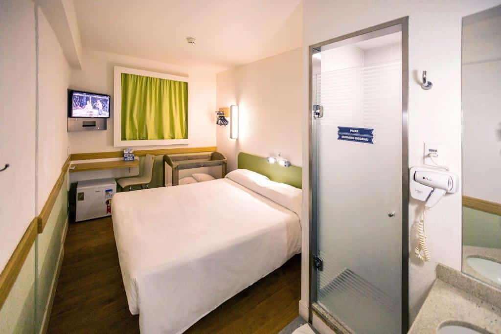 Quarto do hotel com uma cama de casal, um berço pequeno, uma mesinha com uma cadeira, um frigobar, uma tv na parede e uma porta que dá para o banheiro.