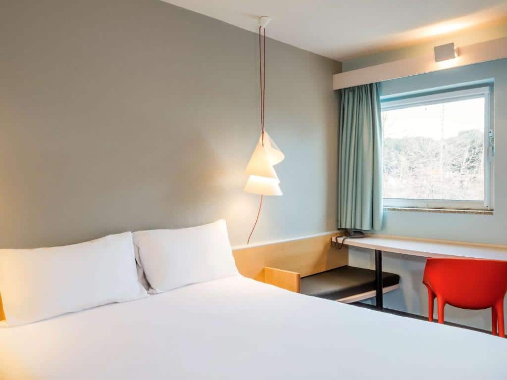 Quarto do Hotel ibis Porto Sao Joao com cama de casal do lado esquerdo da imagem e do lado da cama uma mesa de trabalho com cadeira laranja. Representa hotéis Mercure no Porto.
