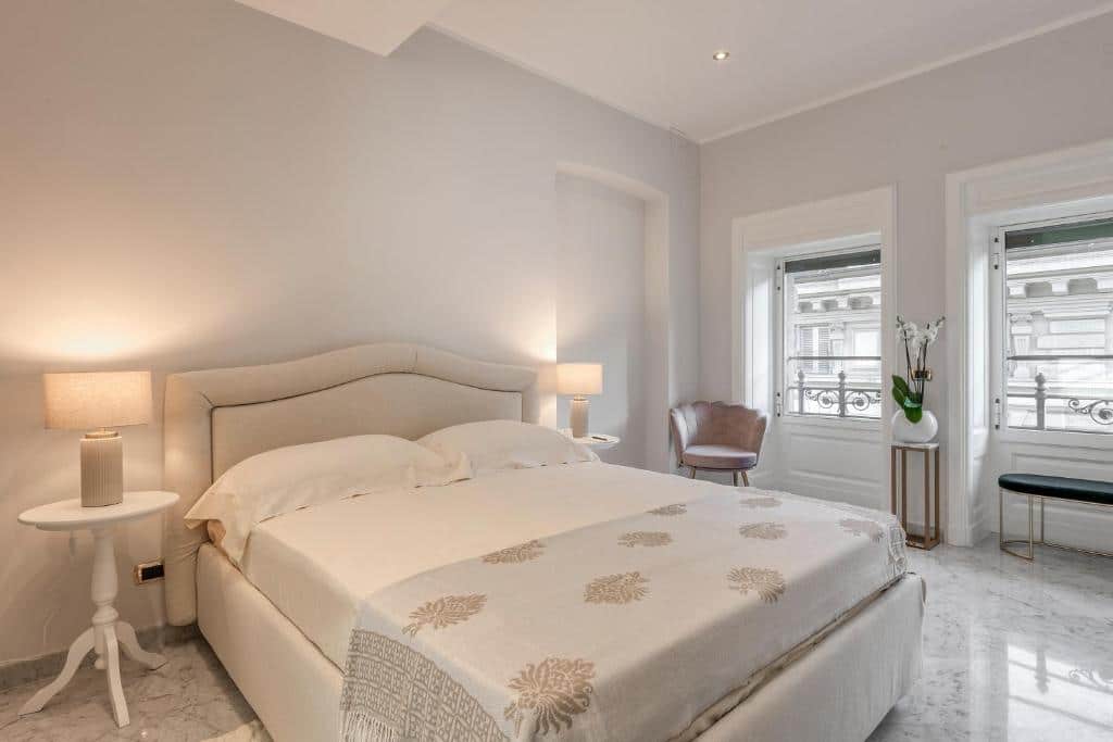 Quarto do Imperiale Suites Milano com duas janelas, uma cama de casal, duas mesinhas pequenas com abajures, além de uma poltrona e um vaso de flor, para representar airbnb em Milão