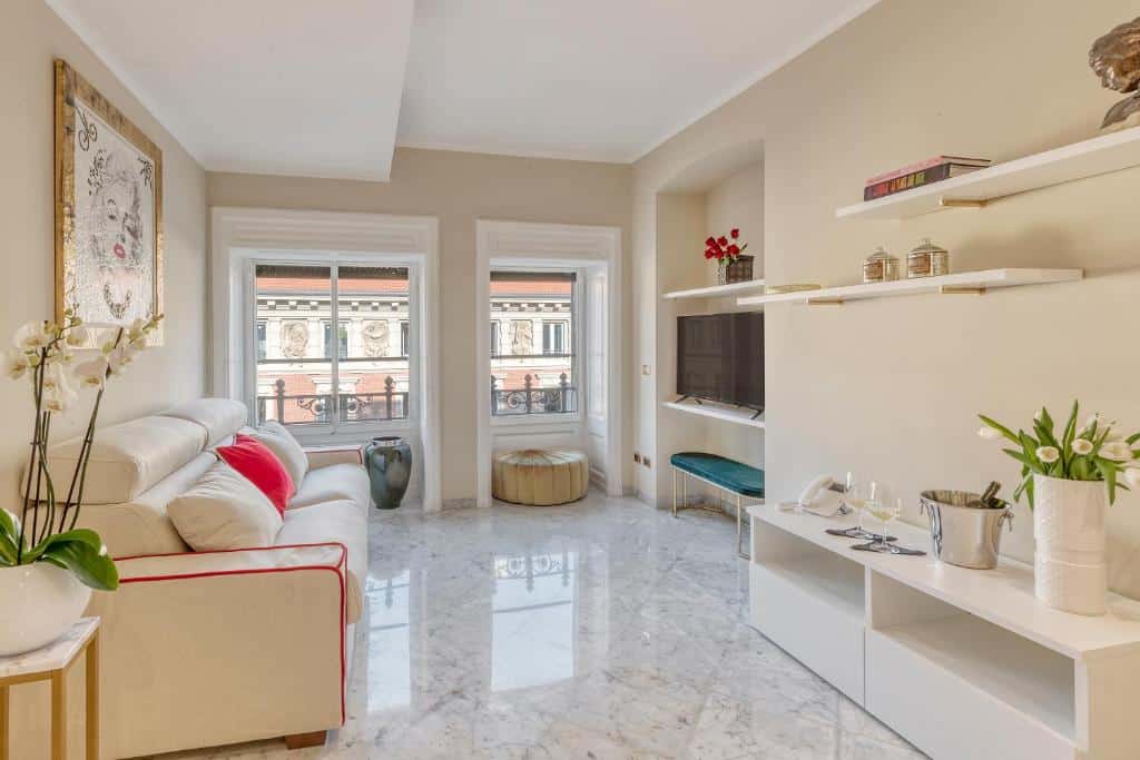 Sala de estar do Imperiale Suites Milano com duas janelas, um sofá com almofadas, uma televisão, diversas prateleiras e um móvel com vasos de plantas, para representar airbnb em Milão
