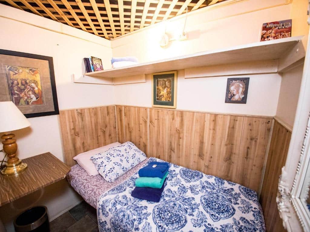 quarto individual do Interfaith Retreats, um dos hostels em Nova York, com uma cama de solteiro no canto, alguns quadros pequenos decorativos presos à parede e uma prateleira alta, sobre a cama.
