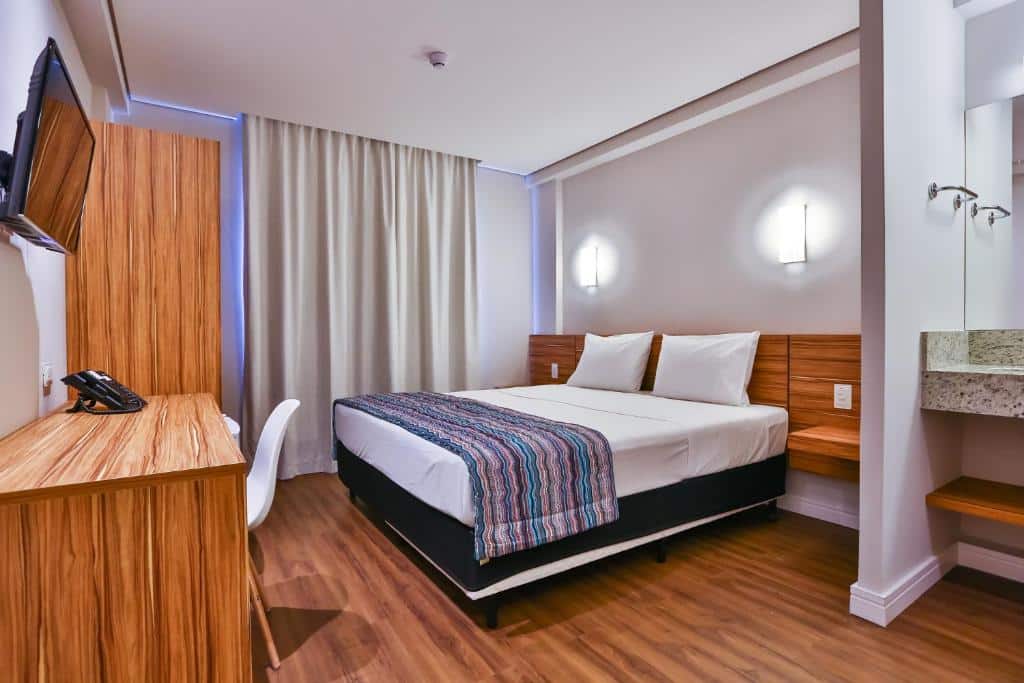 Quarto do hotel com uma cama de casal, chão e móveis de madeira, paredes brancas, tv na parede e uma cortina.