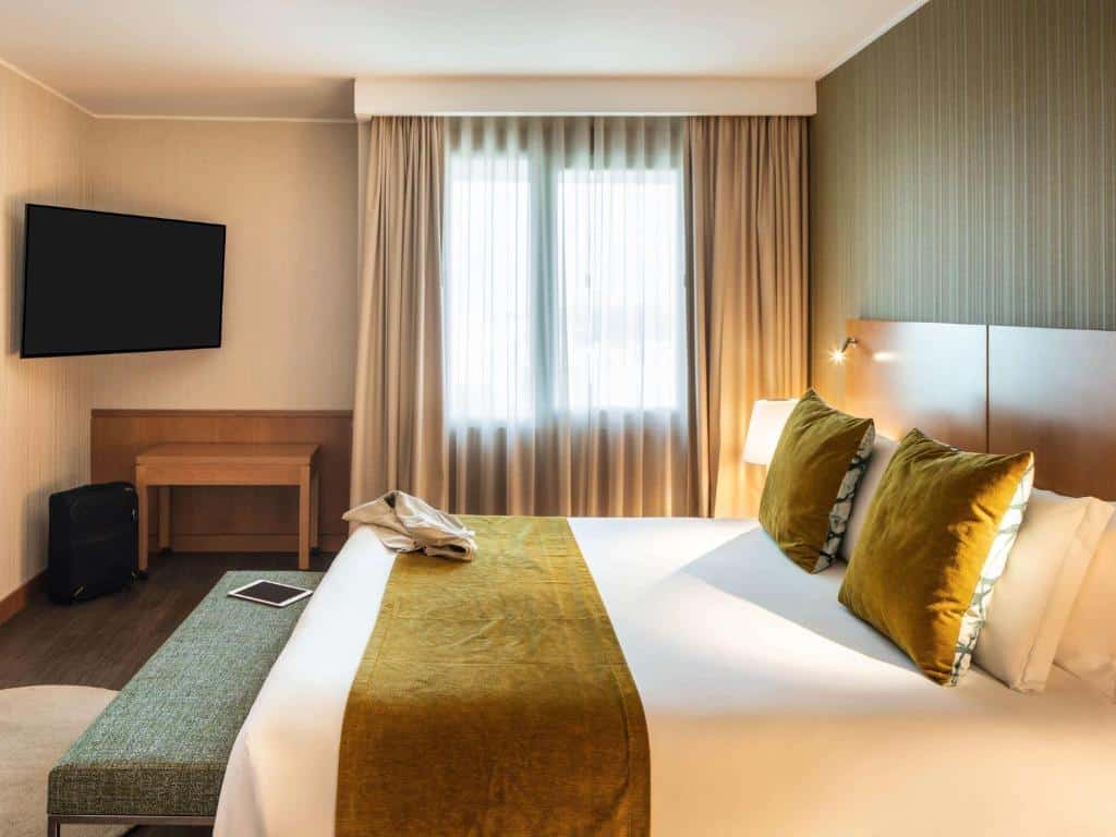 Quarto do Hotel Mercure Porto Gaia com cama de casal do lado direito da imagem, um banco no é da cama e do lado esquerdo da cama uma TV presa na parede. Representa hotéis Mercure no Porto.