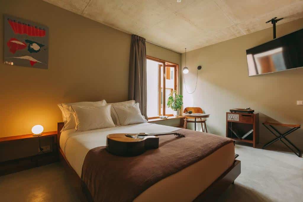 Quarto do  Mouco Hotel – Stay, Listen & Play com cama de casal do lado esquerdo com um violão em cima da cama, do lado esquerdo da cama uma mesa com cadeira e um TV pendurada na parede. Representa hotéis boutique no Porto.