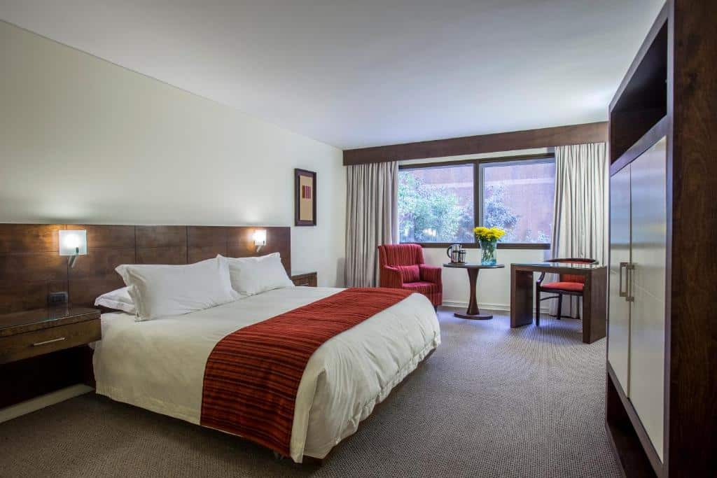 Quarto do MR Hotel Providencia com cama de casal do lado esquerdo da imagem, do lado esquerdo da cama uma poltrona e uma mesa com cadeira. Representa hotéis em Providencia em Santiago.