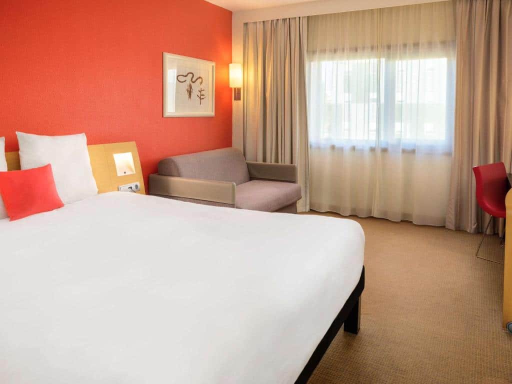 Quarto do Novotel Porto Gaia com cama de casal do lado esquerdo da imagem e do lado esquerdo da cama uma poltrona. Representa hotéis Mercure no Porto.