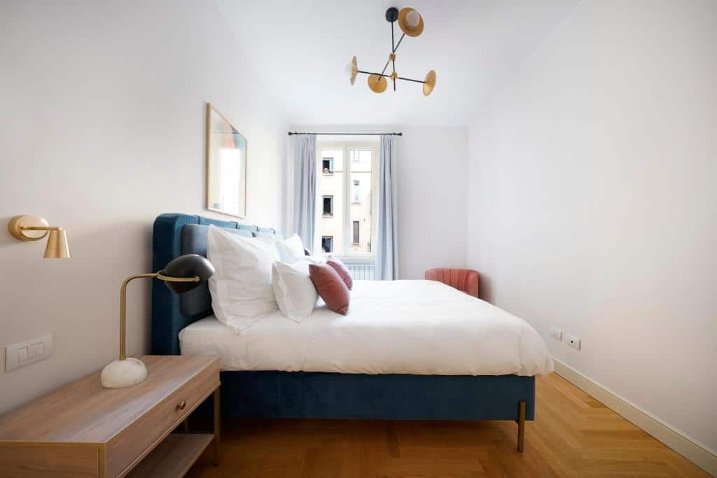 Quarto do numa I Loreto Apartments com uma cama de casal, piso que imita madeira, uma janela com cortinas, uma mesinha de cabeceira com uma luminária e um lustre com diversas lâmpadas