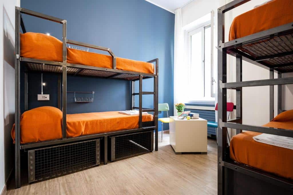 Quarto do Ostello Bello Grande com duas beliches pretas com roupas de cama laranja, no canto do quarto, embaixo da janela, há uma pequena mesinha com duas cadeiras, para representar hostels em Milão