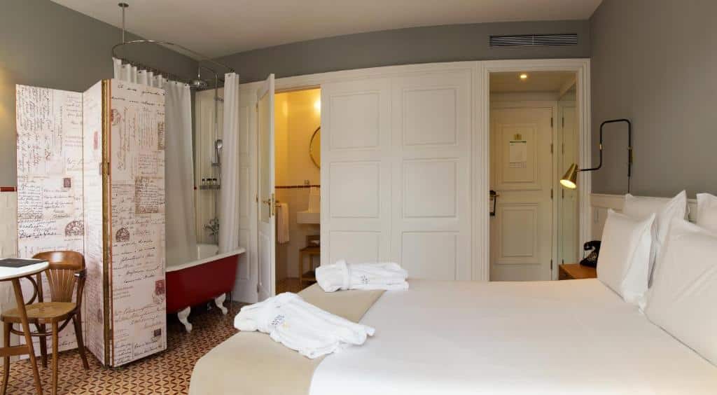Quarto do Porto A.S. 1829 Hotel com cama de casal do lado direito da imagem, do lado esquerdo da cama uma cômoda com telefone e em frente a cama uma banheira. Representa hotéis bem localizados no Porto.