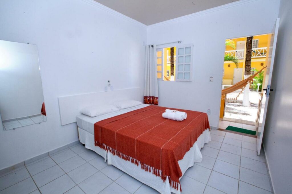 Quarto da Pousada Barra Bonita com uma cama de casal com duas toalhas em cima dela, um espelho, uma janela e uma porta aberta que mostra uma rede de descanso no lado de fora. Foto para ilustrar post sobre pousadas em Barra de São Miguel.