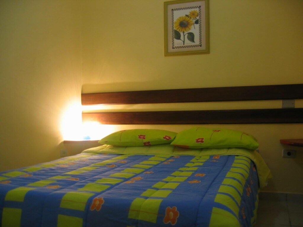 Quarto da Pousada Boramar. Uma cama de casal com dois travesseiros. Do lado esquerdo uma luminária.