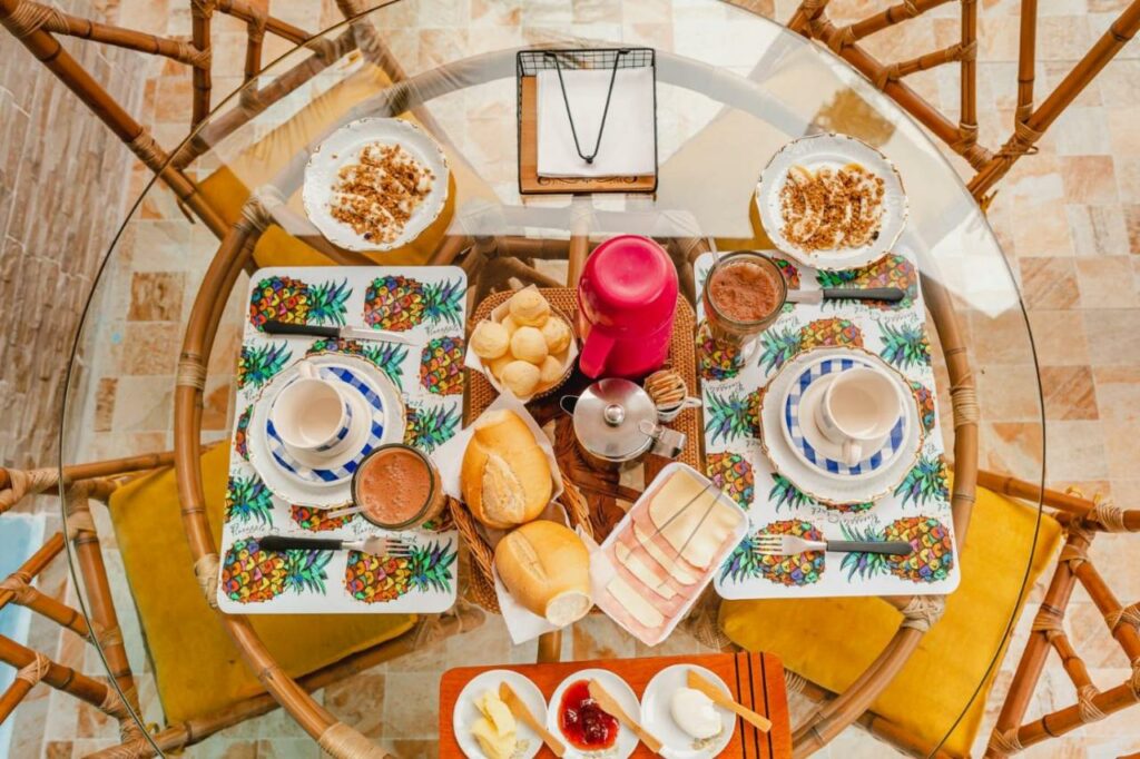 Café da manhã da Pousada Oásis Café. Uma mesa redonda, de vidro e quatro cadeiras. Uma mesa cheia de variedade do café da manhã, com pães, sucos, xícaras, frios e frutas.