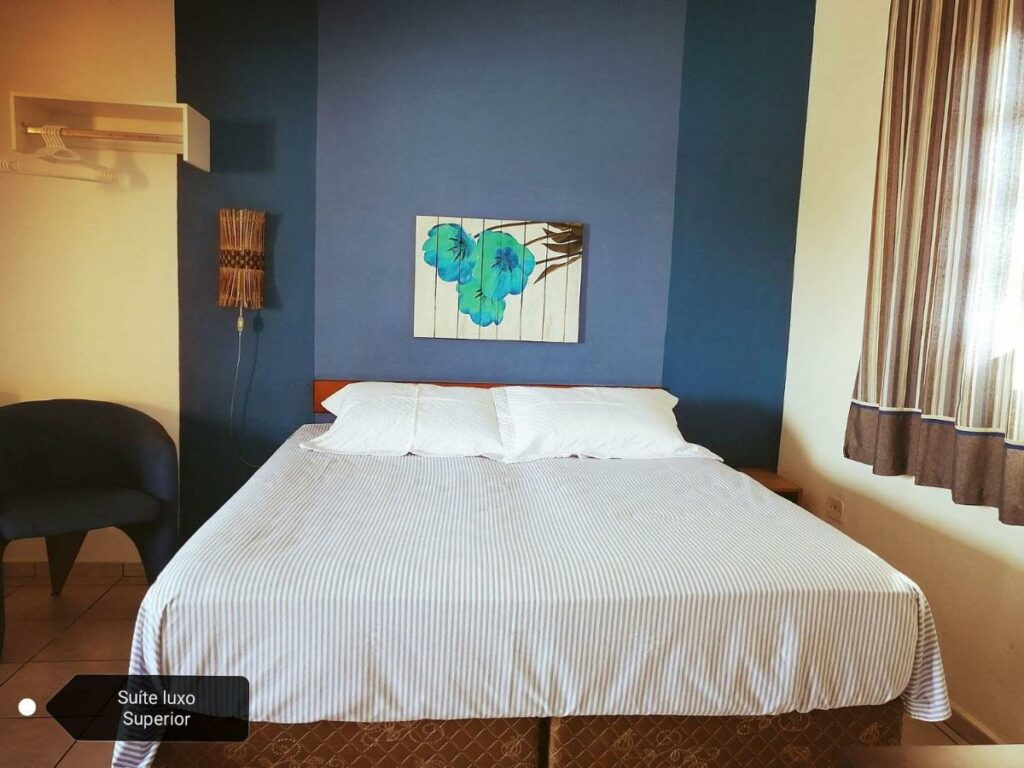 Uma cama de casal na Pousada Pier36. Do lado esquerdo uma poltrona e do lado direito a janela do quarto.
