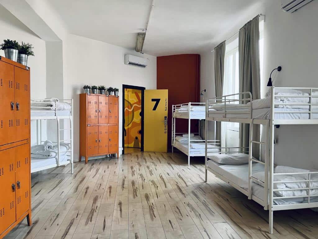 Quarto do QUO Milano com três beliches, armários com chave na cor laranja, há uma ampla janela com cortinas e ar-condicionado de teto