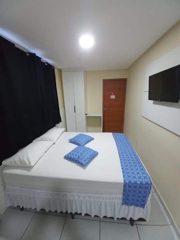 Quarto de hotel simples com cama de casal com colcha branca e detalhes em azul, TV em frente a cama, pequeno armário branco ao lado da cama e porta de madeira. Imagem para ilustrar o post hotéis em Campina Grande.