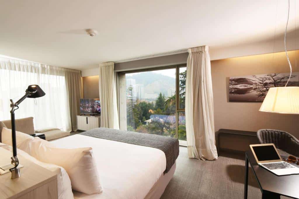 Quarto do Solace Hotel Santiago com cama de casal no lado esquerdo da imagem no centro e em frente a cama uma janela ampla com vista para a cidade. Representa hotéis em Providencia em Santiago.