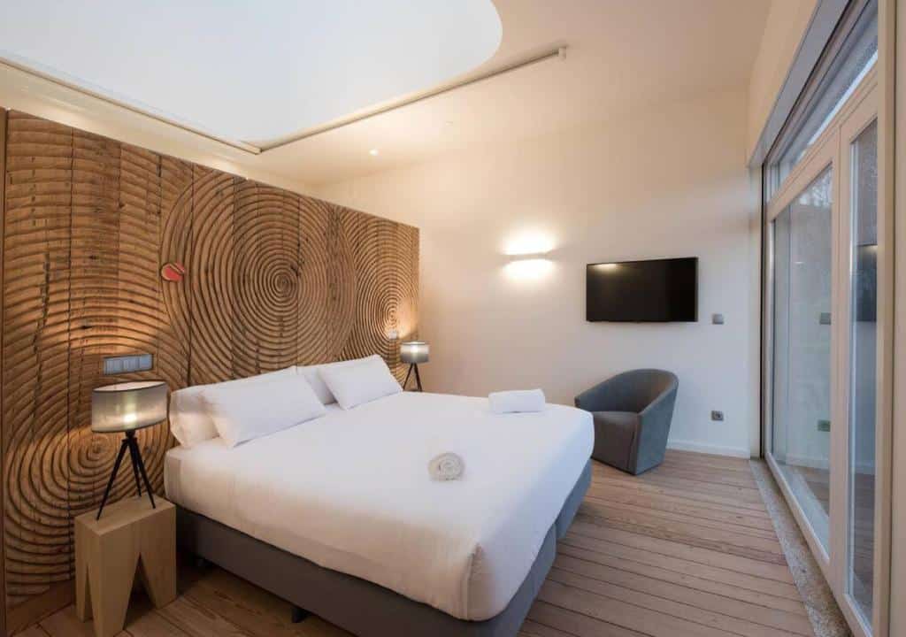 Quarto privado do The Central House Porto Ribeira com cama de casal do lado esquerdo, duas cômodas ao lado da cama com luminária e uma poltrona cinza do lado esquerdo da imagem.