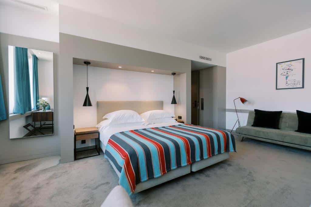Quarto do The Editory Artist Baixa Porto Hotel com cama de casal do lado esquerdo com duas comodas ao lado da cama, do lado direito da cama um sofá de dois lugares e do lado esquerdo um espelho comprido na parede. Representa hotéis boutique no Porto.