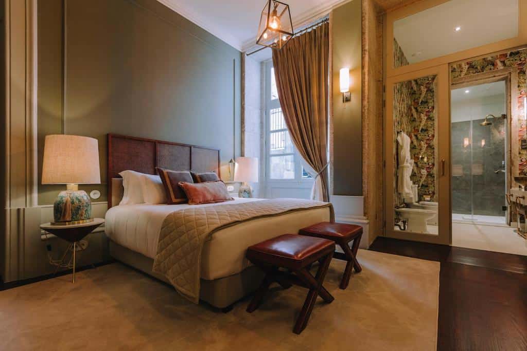 Quarto do Torel 1884 Suites & Apartments com cama de casal do lado esquerdo, duas comodas com luminária em cada lado da cama e ao pé da cama dois bancos.