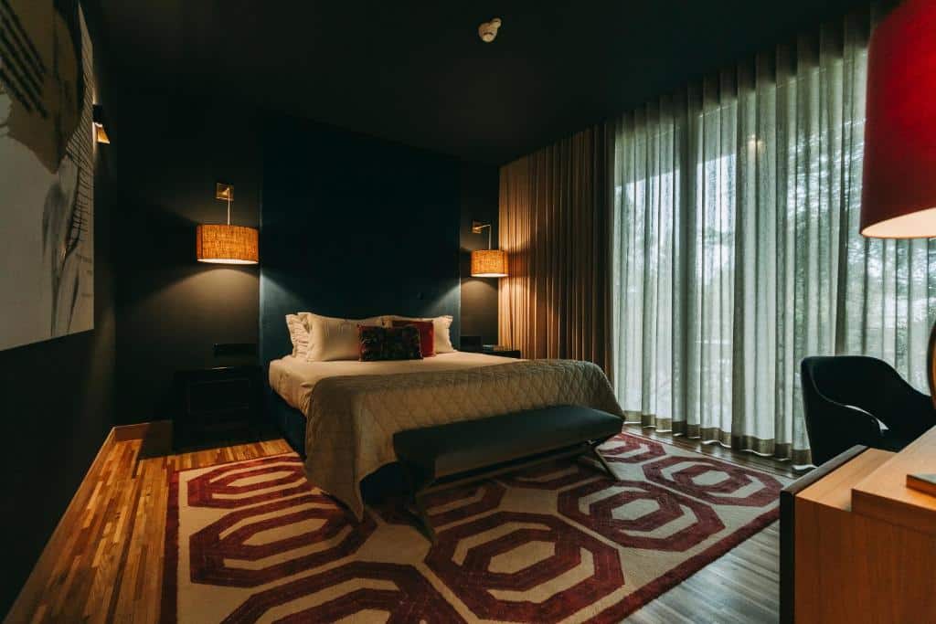 Quarto do Torel Avantgarde com cama de casal do lado esquerdo da imagem no centro com duas cômodas ao lado da cama e em frente a cama uma mesa de trabalho.  Representa hotéis bem localizados no Porto.